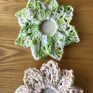 May include: Two crocheted flower hair scrunchies. One is green and white, the other is cream with pink and purple accents.