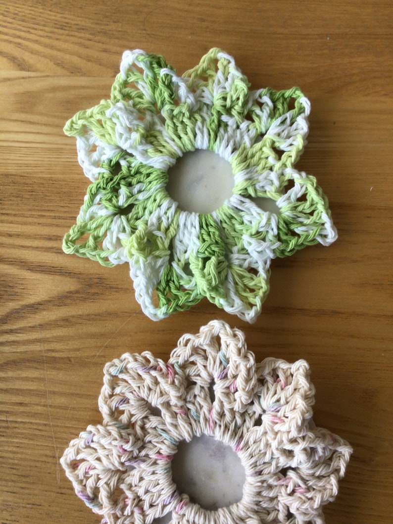 May include: Two crocheted flower hair scrunchies. One is green and white, the other is cream with pink and purple accents.