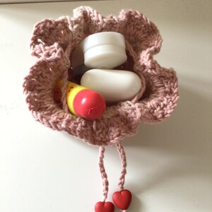 May include: A pink crocheted drawstring bag with two red heart-shaped charms. The bag is filled with small items, including a white round container, a white oval-shaped soap, and a yellow and red lip balm.