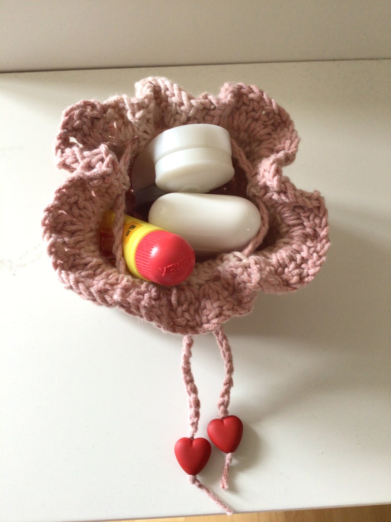 May include: A pink crocheted drawstring bag with two red heart-shaped charms. The bag is filled with small items, including a white round container, a white oval-shaped soap, and a yellow and red lip balm.
