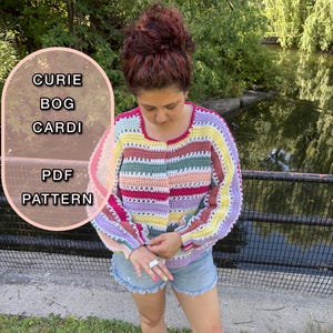 May include: A multi-coloured, striped crochet cardigan with the text "CURIE BOG CARDI PDF PATTERN". The cardigan has a square neckline and long sleeves. The colour scheme includes pink, yellow, green, and purple stripes. The image is taken outdoors.