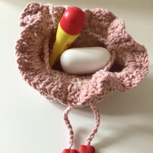 May include: A pink crocheted drawstring bag with two red heart-shaped beads on the cords. The bag contains a yellow and red marker and a white oval-shaped object.