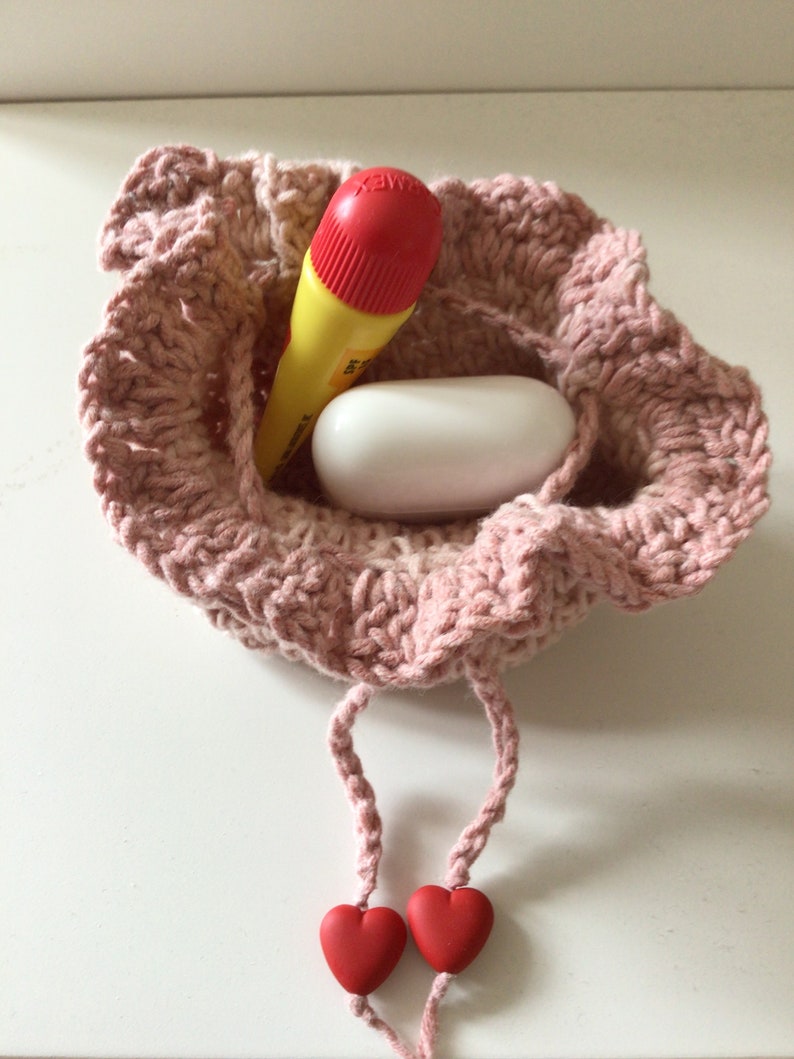 May include: A pink crocheted drawstring bag with two red heart-shaped beads on the cords. The bag contains a yellow and red marker and a white oval-shaped object.