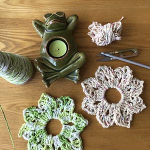 May include: Two crocheted flower coasters, one in green and white, the other in beige with pink and blue accents. A ceramic frog candle holder with a green glaze and a tealight candle inside. A ball of yarn, a pair of scissors, and a crochet hook are also pictured.