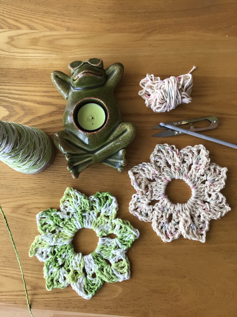 May include: Two crocheted flower coasters, one in green and white, the other in beige with pink and blue accents. A ceramic frog candle holder with a green glaze and a tealight candle inside. A ball of yarn, a pair of scissors, and a crochet hook are also pictured.