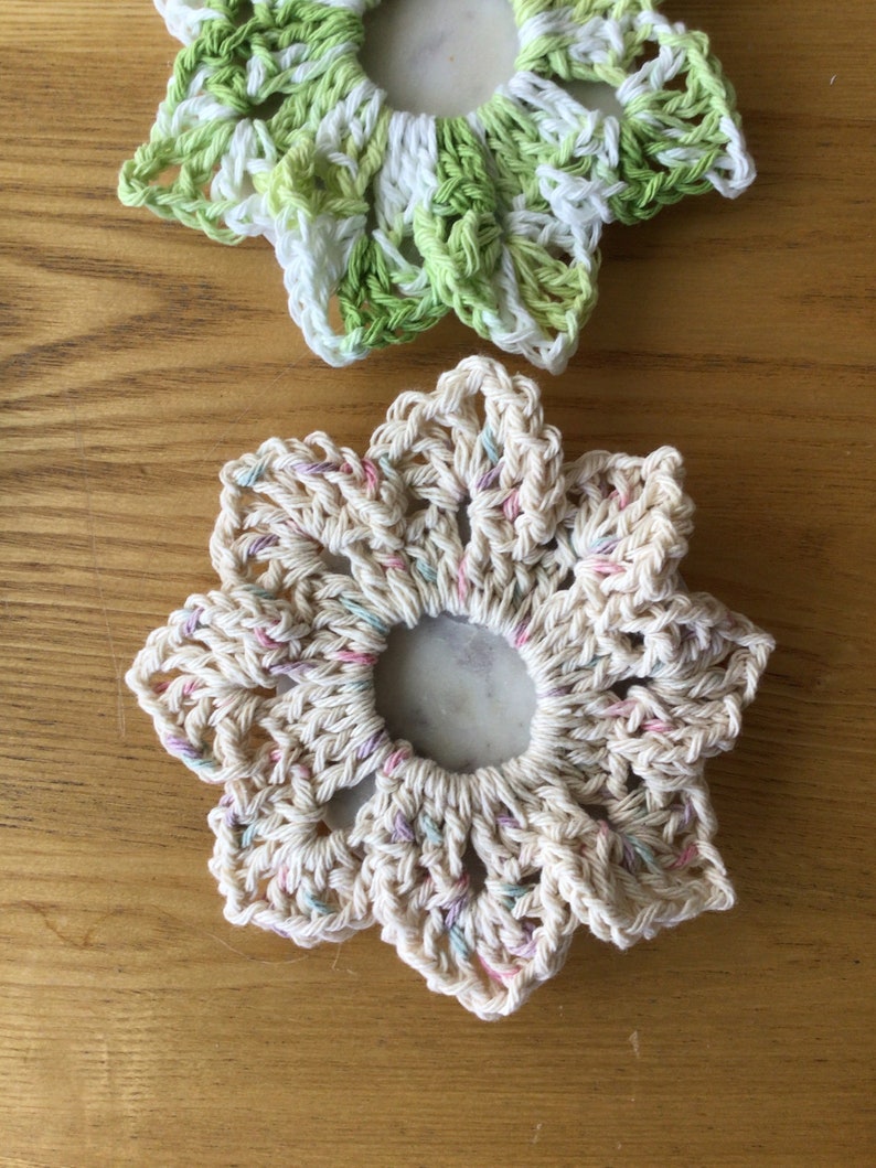 May include: Two crocheted flower-shaped coasters. One is white with pink and blue accents, the other is green and white.