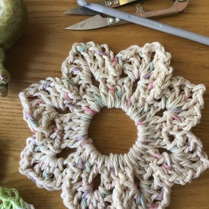 May include: A crocheted flower-shaped coaster in cream with pink, green, and purple accents. The coaster is made with a cotton yarn and has a hole in the center.