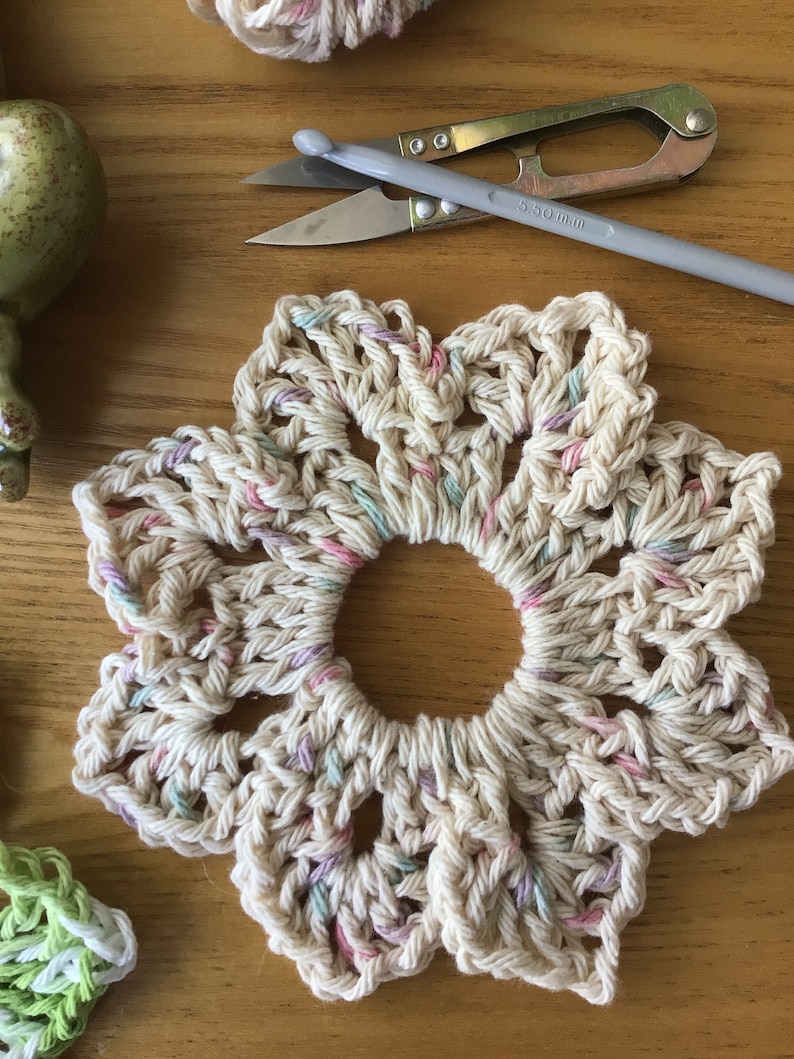 May include: A crocheted flower-shaped coaster in cream with pink, green, and purple accents. The coaster is made with a cotton yarn and has a hole in the center.