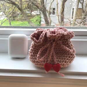 May include: A pink crocheted drawstring bag with red heart-shaped embellishments. The bag is sitting on a white windowsill.