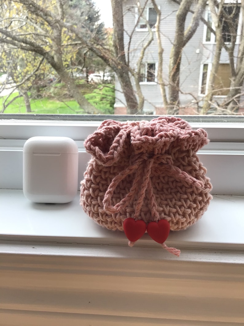 May include: A pink crocheted drawstring bag with red heart-shaped embellishments. The bag is sitting on a white windowsill.