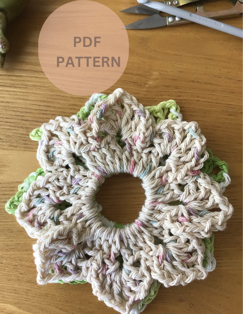 May include: A crocheted flower-shaped coaster in cream, green, and pink. The coaster is made with a chunky yarn and has a hole in the center. The image includes the text "PDF PATTERN".