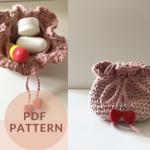 May include: A pink crocheted drawstring bag with two red heart-shaped beads on the drawstring. The bag is empty and sitting on a white surface. The text "PDF PATTERN" is printed on a pink background.