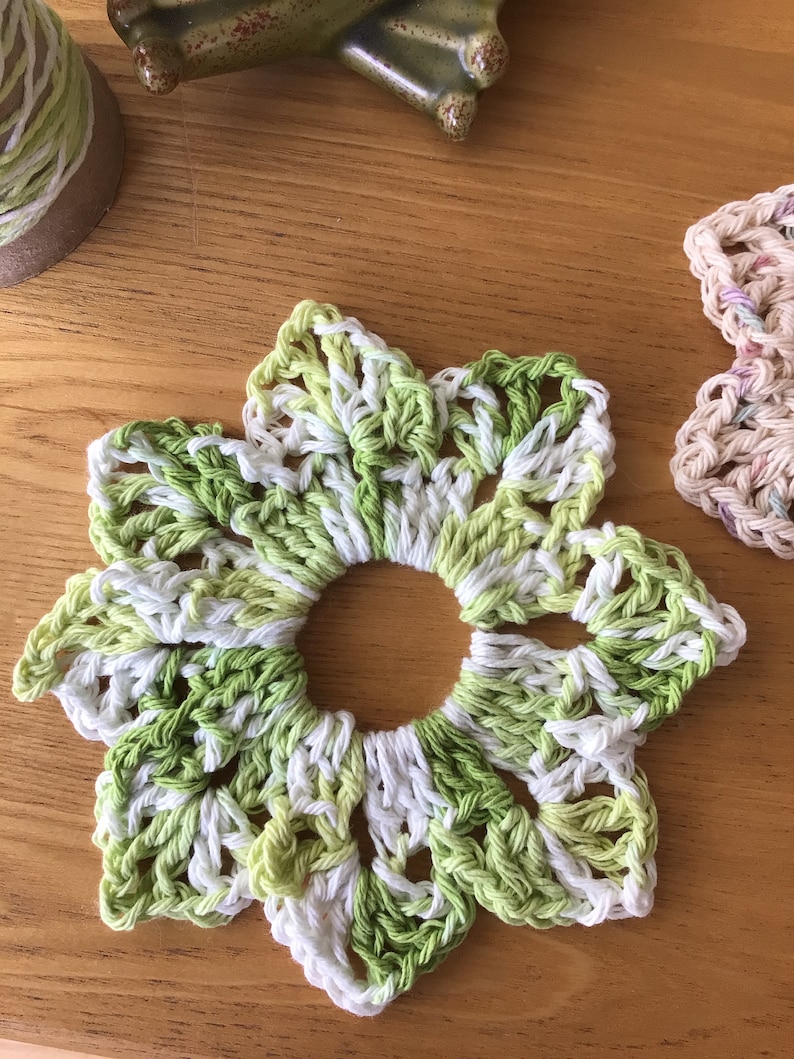 May include: A crocheted flower coaster in shades of green and white. The coaster is made with a delicate, lacy pattern and has a circular hole in the center.