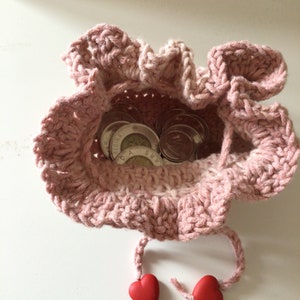 May include: A pink crocheted drawstring bag with a ruffled edge, containing several Canadian coins. The bag has two red heart-shaped beads attached to the drawstrings.