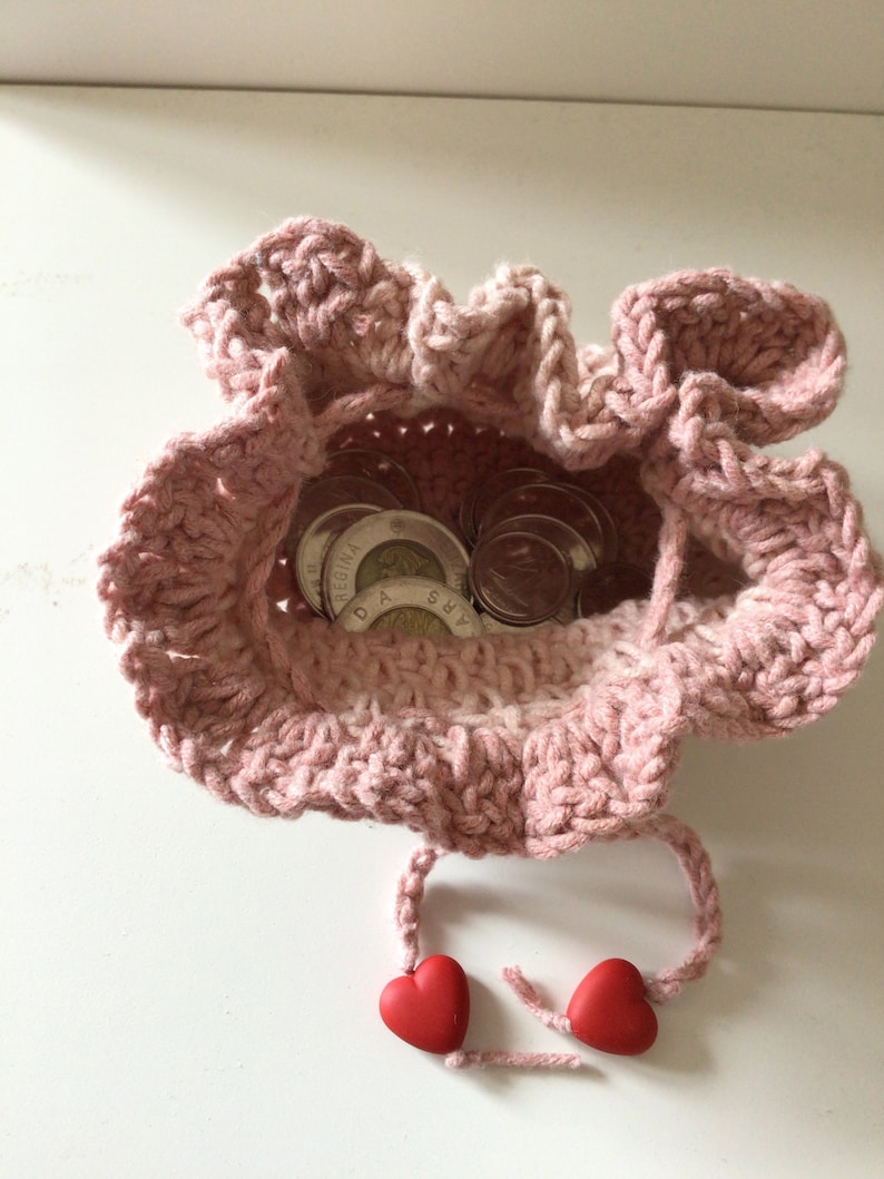 May include: A pink crocheted drawstring bag with a ruffled edge, containing several Canadian coins. The bag has two red heart-shaped beads attached to the drawstrings.
