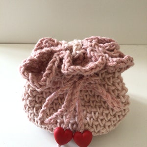 May include: A pink crocheted drawstring bag with two red heart-shaped buttons. The bag is made with a textured stitch and has a ruffled top.