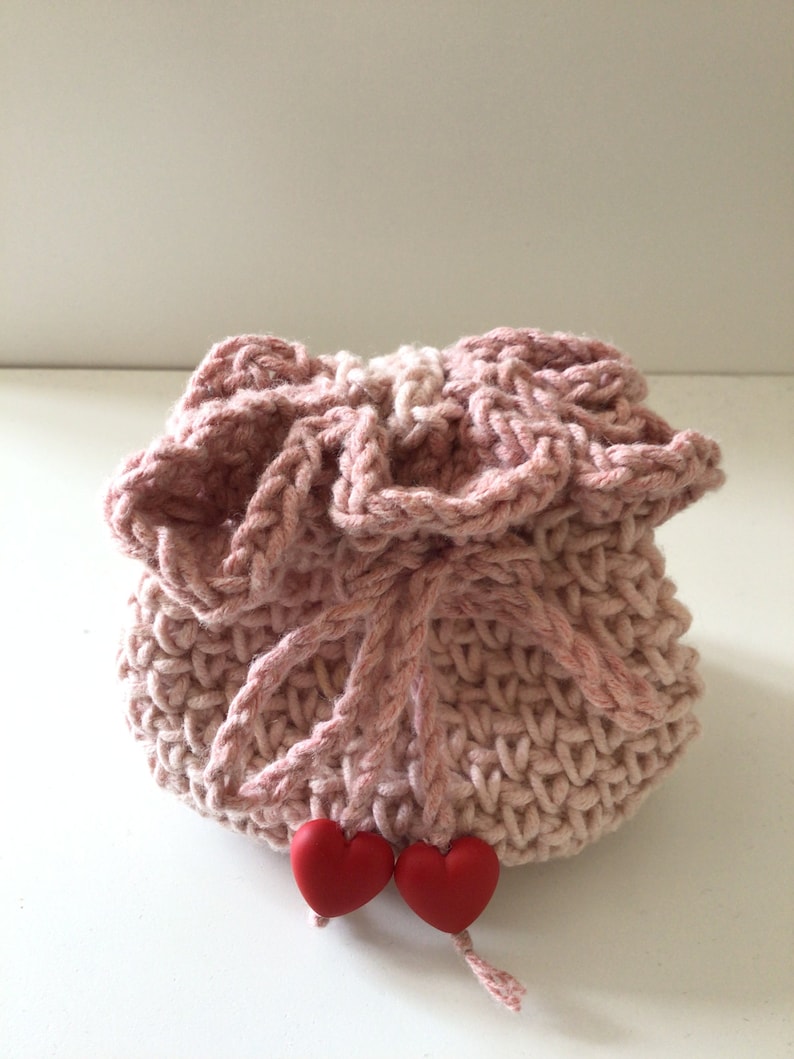 May include: A pink crocheted drawstring bag with two red heart-shaped buttons. The bag is made with a textured stitch and has a ruffled top.