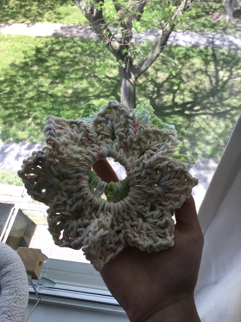 May include: A crocheted flower-shaped hair scrunchie in white with pink, green, and light blue accents.