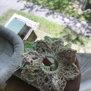 May include: A crocheted flower-shaped hair scrunchie in cream, green, and pink. The scrunchie is made with a variety of stitches and has a delicate, feminine look.