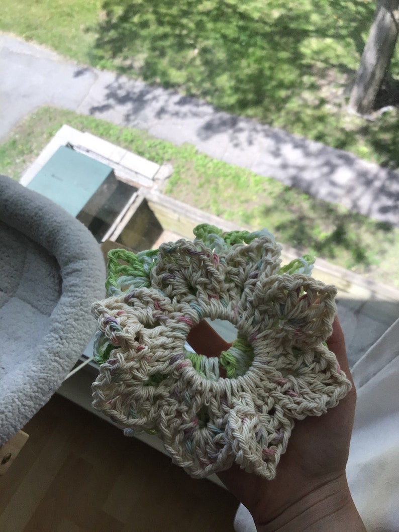 May include: A crocheted flower-shaped hair scrunchie in cream, green, and pink. The scrunchie is made with a variety of stitches and has a delicate, feminine look.
