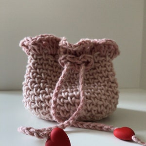 May include: A pink crocheted drawstring bag with a ruffled top and two red heart-shaped charms attached to the drawstring.