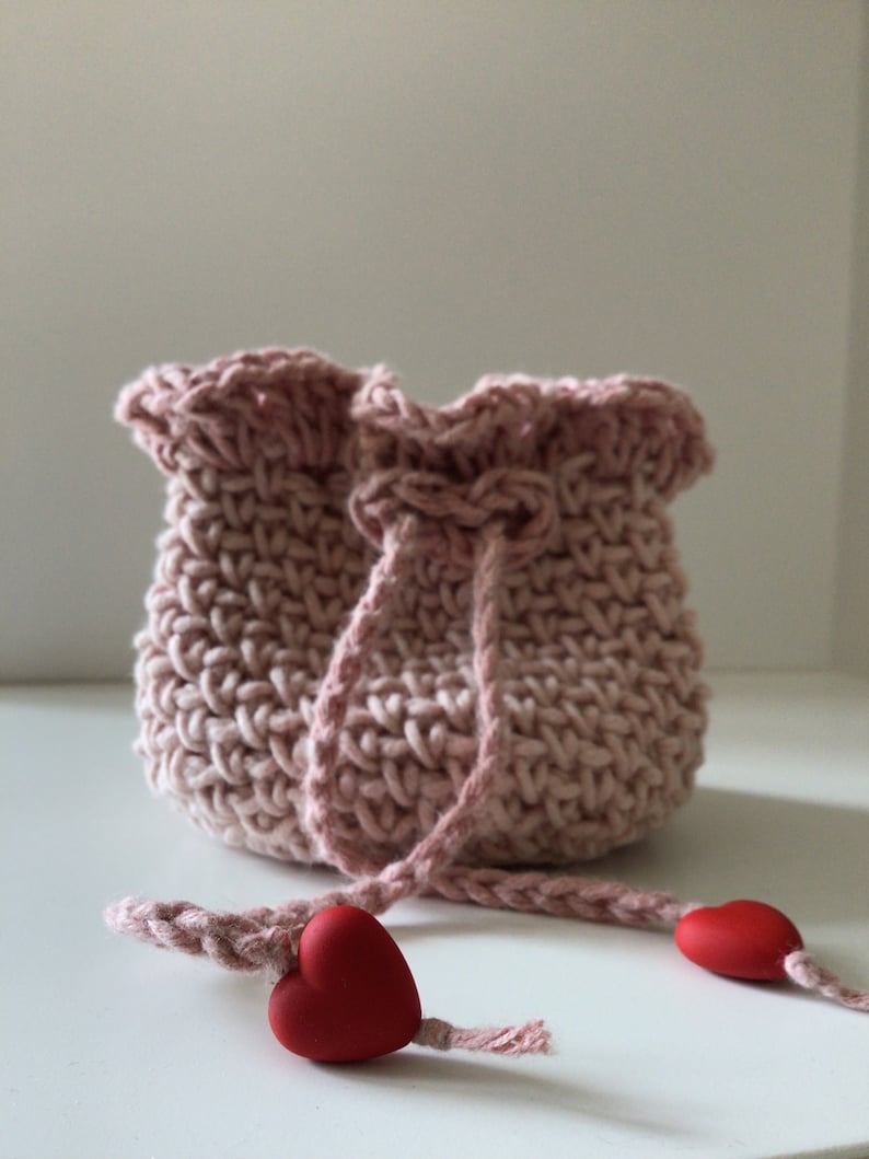 May include: A pink crocheted drawstring bag with a ruffled top and two red heart-shaped charms attached to the drawstring.