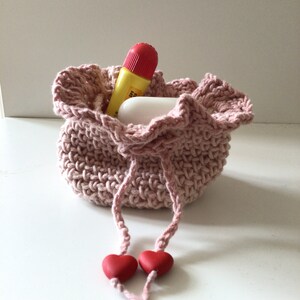 May include: A pink crocheted drawstring bag with two red heart-shaped beads on the ends of the drawstrings. The bag is filled with a yellow and red tube of lip balm and a white bar of soap.