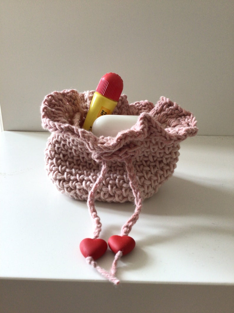 May include: A pink crocheted drawstring bag with two red heart-shaped beads on the ends of the drawstrings. The bag is filled with a yellow and red tube of lip balm and a white bar of soap.
