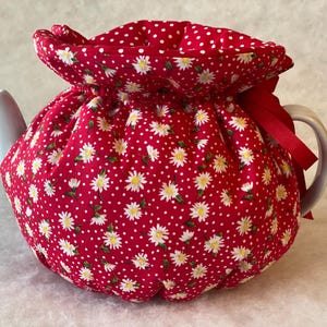 Könnte beinhalten: Rote Teekanne-Kuscheldecke mit einem Muster aus weißen Gänseblümchen und Punkten. Die Kuscheldecke hat einen Kordelverschluss und ein rotes Band.