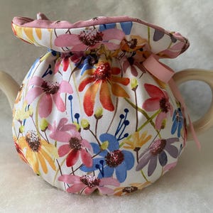 Puede incluir: Una tetera de cerámica con un cubre tetera floral. El cubre tetera es blanco con un estampado floral colorido en rosa, naranja, amarillo y azul. Una cinta rosa está atada al asa. La tetera tiene un asa y un pico de color beige claro.