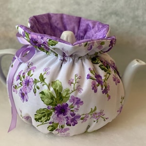 May include: A white teapot with a floral tea cozy. The cozy features a purple floral pattern with green leaves and a purple interior. A purple ribbon is tied around the teapot. The teapot has a white handle and spout.