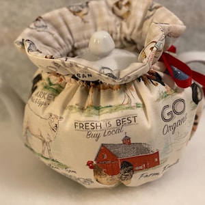 Könnte beinhalten: Eine weiße Keramik-Teekanne mit einer Farm-Themen-Hülle. Die Hülle ist cremefarben mit Illustrationen von Nutztieren, einer roten Scheune und dem Text "FRESH IS BEST Buy Local" und "GO Organic". Ein rotes Band akzentuiert die Hülle.