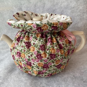 May include: A cream-colored teapot with a floral tea cozy. The cozy is made of fabric with a pattern of burgundy, yellow, and pink roses on a white background. A pink ribbon is tied to the handle. The lid of the cozy has a button.