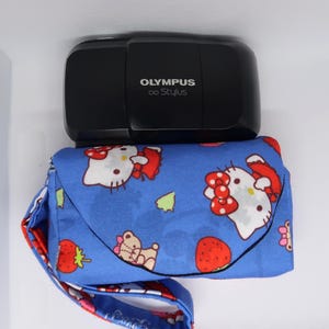 May include: A black Olympus oo Stylus camera rests atop a blue fabric case. The case features a pattern of white cartoon cat faces with red bows, strawberries, and small teddy bears. The case has a matching strap.