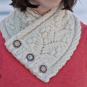 May include: A white knitted cowl neck scarf with a cable knit pattern and decorative buttons. The scarf is worn by a person with long brown hair.