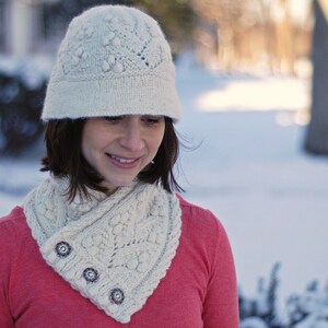 May include: A woman wearing a white knitted hat and a white knitted scarf with a pink sweater. The hat has a cable knit pattern and the scarf has a lace pattern.