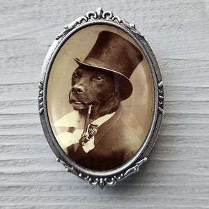 May include: Oval-shaped, silver-toned brooch featuring a sepia-toned portrait of a dog wearing a top hat and smoking a pipe. The dog is dressed in a suit and tie, giving the brooch a vintage, whimsical feel.