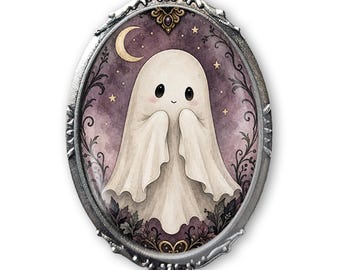 May include: An oval brooch featuring a silver-toned ornate metal frame surrounding a central illustration of a small, white, cartoon-style ghost. The background is a deep purple gradient with a crescent moon, small stars, and dark foliage accents.