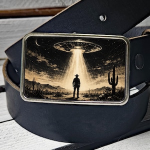 May include: A black leather belt featuring a rectangular metal buckle with a graphic print of a desert landscape at night. The image shows a silhouette of a person in a cowboy hat standing under a bright beam of light from a flying saucer.