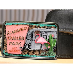 May include: A silver belt buckle with a pink flamingo, a vintage trailer, and a sign that reads "Flamingo Trailer Park No Vacancy" on a turquoise and leopard print background.
