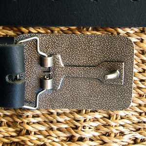 May include: A silver metal belt buckle with a textured surface and a hook closure. The buckle is attached to a black leather belt.