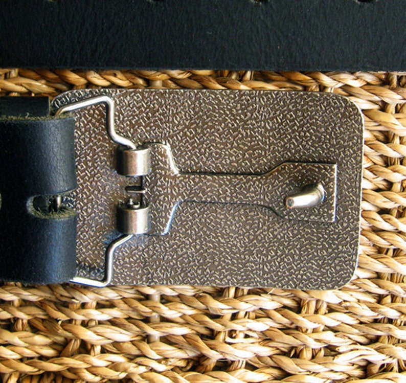 May include: A silver metal belt buckle with a textured surface and a hook closure. The buckle is attached to a black leather belt.