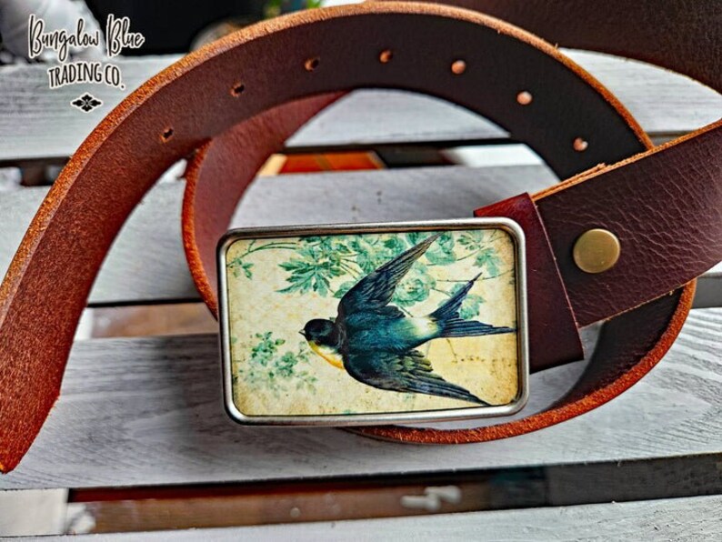 May include: A brown leather belt with a silver buckle featuring a vintage image of a blue bird on a floral background. The buckle has the text "Bungalow Blue Trading Co."