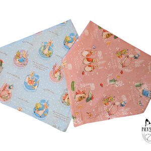 Può includere: Due fazzoletti quadrati a tema Peter Rabbit. Uno è azzurro con pois bianchi e illustrazioni di Peter Rabbit. L'altro è rosa con illustrazioni di Peter Rabbit e dettagli floreali.