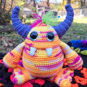 May include: A crocheted monster toy with a striped orange, pink, and yellow body. The monster has large blue ears, green hair, and large blue eyes.