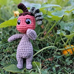 May include: A crocheted amigurumi toy with a red head, grey body, and green leafy hair. The toy is standing on a bed of green grass.