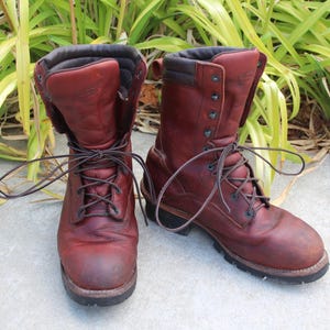 May include: A pair of dark reddish-brown leather work boots featuring a high-top design, dark brown padded collars, black metal eyelets, and dark brown laces. The boots have a rounded toe, a thick black rubber sole, and visible stitching.