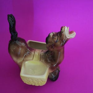 May include: A ceramic donkey planter in shades of brown and tan, with a black tail and open mouth. The donkey has a basket-shaped container on its back, perfect for small plants or flowers. The planter is set against a vibrant purple background.