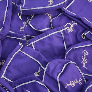 May include: A pile of purple drawstring bags with white stitching and the words "Crown Royal" embroidered in gold.