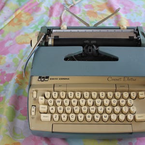May include: A vintage SCM Smith-Corona Coronet Electric typewriter in a light blue and beige color scheme. The keyboard features white keys with black lettering. The typewriter sits on a floral patterned fabric.